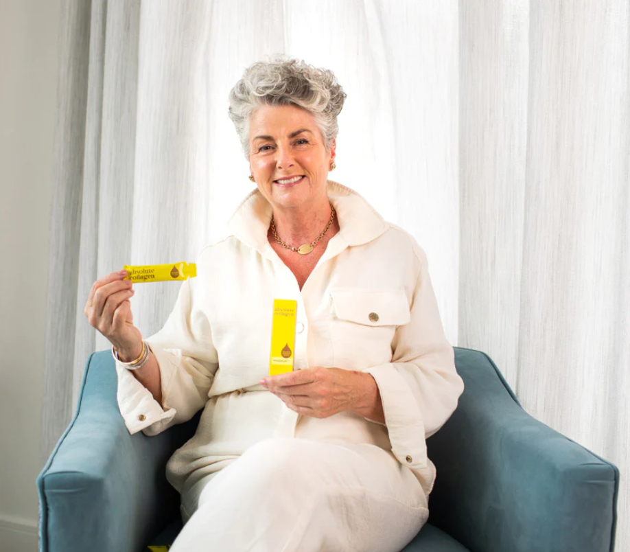 Image of Maxine Laceby, sat on a blue chair, showcasing the yellow collagen supplement sachet and the collagen booster serum