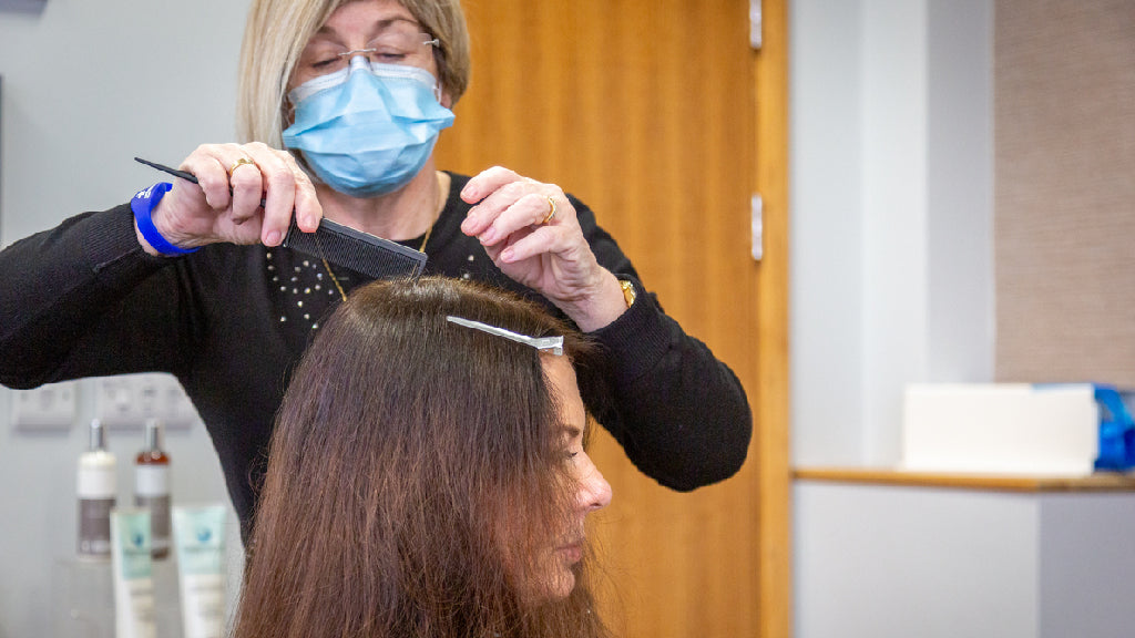 Photo showing Eva Proudman, a white woman with short blonde hair, wearing a blue face mask and studying the hair of a white woman with long brown hair as she sits in a clinical room