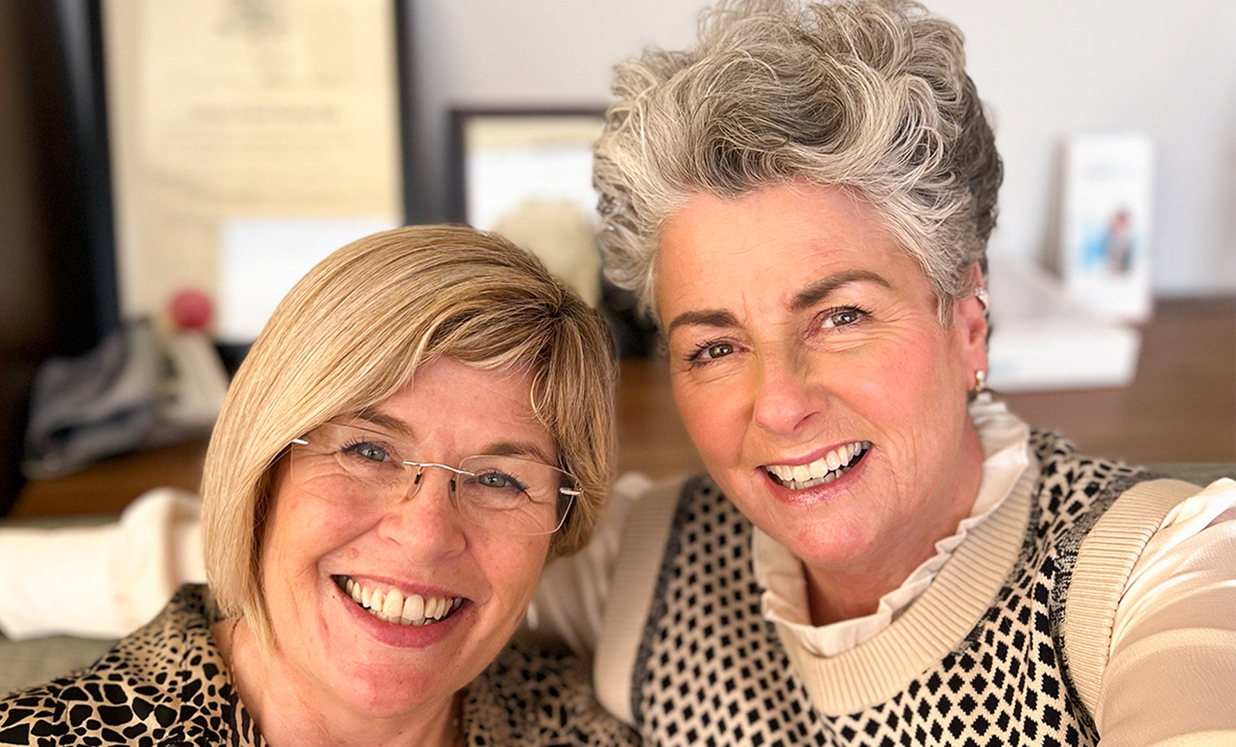 Photo showing two white women smiling at the screen, they are both middle aged with short light hair and wearing earthy colour clothes