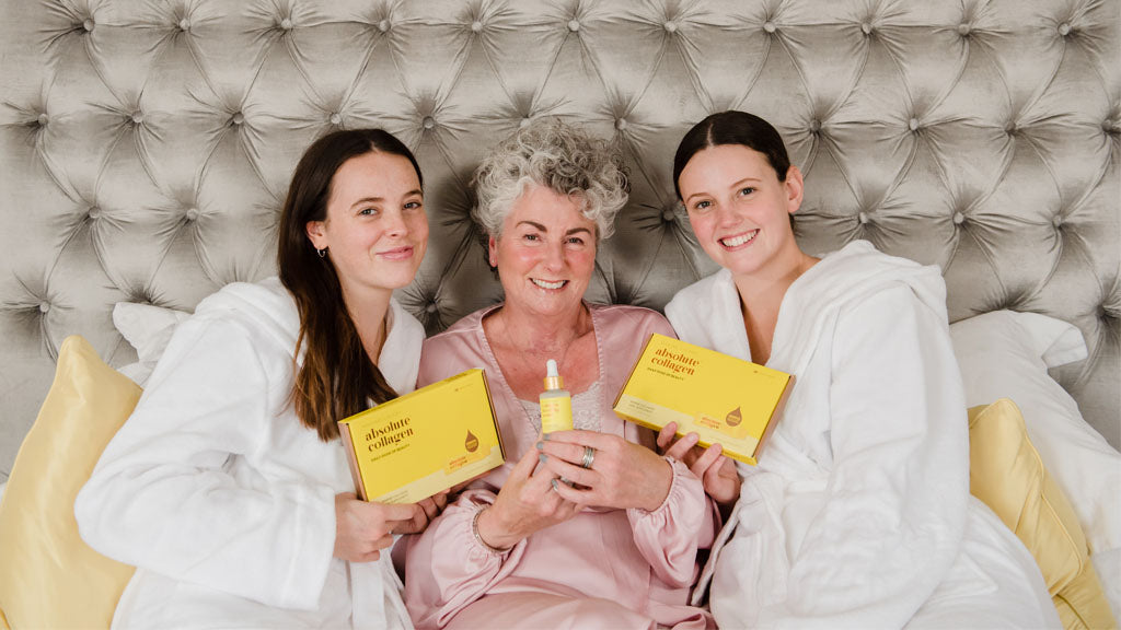 Photo showing Maxine, Darcy and Margot Laceby smiling at the camera, they are wearing dressing gowns and sitting in bed against a grey velvet headboard while holding Absolute Collagen products.